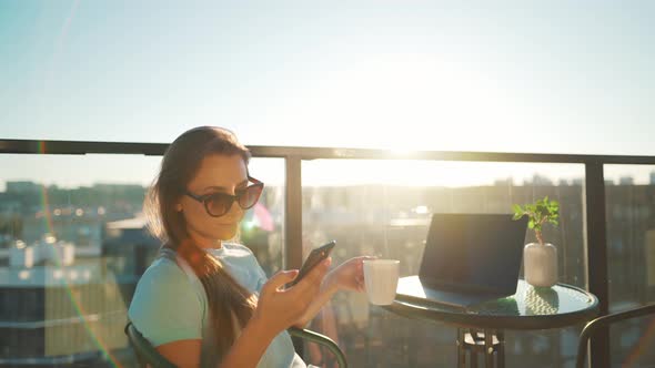 Woman Using Smartphone on the Balcony Against the Backdrop of the Sunset. Outdoor Home Office