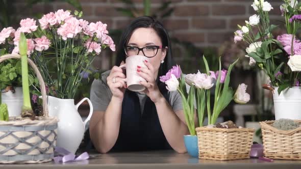Young girl florist drinks coffee among blooming roses, hyacinths and carnations.