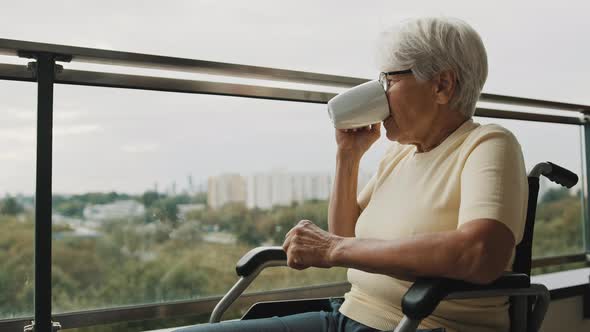 Senior Gray Haired Woman with Eyeglasses Drinking Cup of Coffee in the Wheelchair in Front