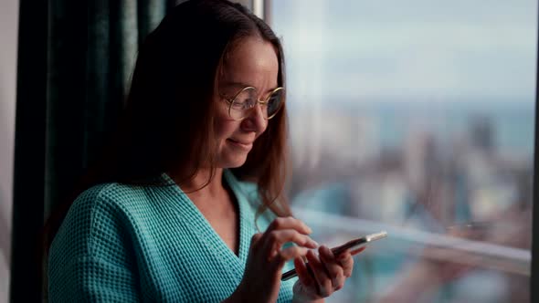 Pretty Adult Woman in Round Glasses is Standing at the Window of Her Apartment in the Morning with