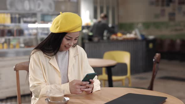 Pleasant Asian Woman Sitting at Table and Using Smartphone