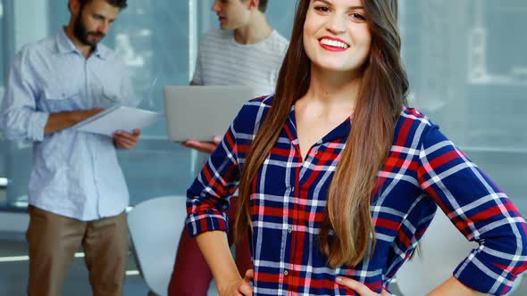 Smiling female executive standing with hands on hip while colleagues discussing in background