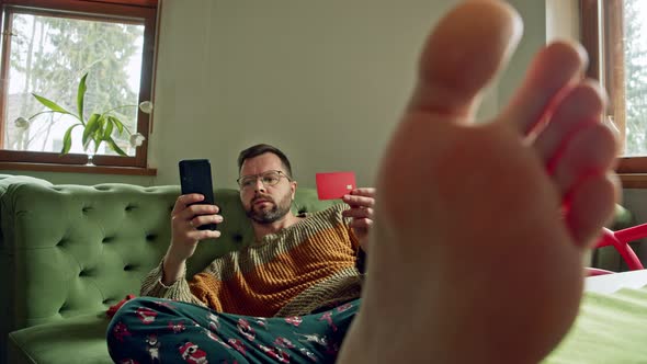 a Bored Man at Home Uses a Credit Card to Pay for Smartphone