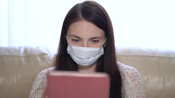 Woman in a Medical Mask Uses a Tablet at Home During Quarantine