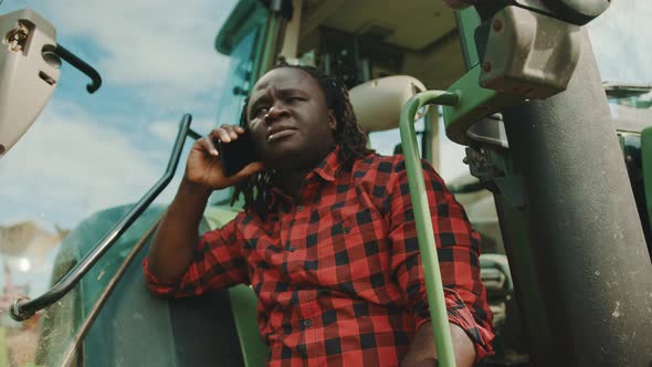 African Farmer Arguing on the Smartphone While Leaning on the Green Tractor