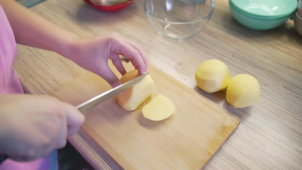 Woman is Finely Slicing Young Fresh Potatoes in the Kitchen with Sharp Knife on Wooden Board for