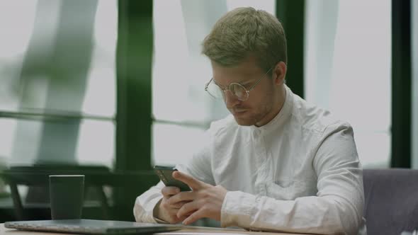 Handsome Man in White Shirt and Glasses is Using Smartphone in Cafe or Coworking Area Businessman or