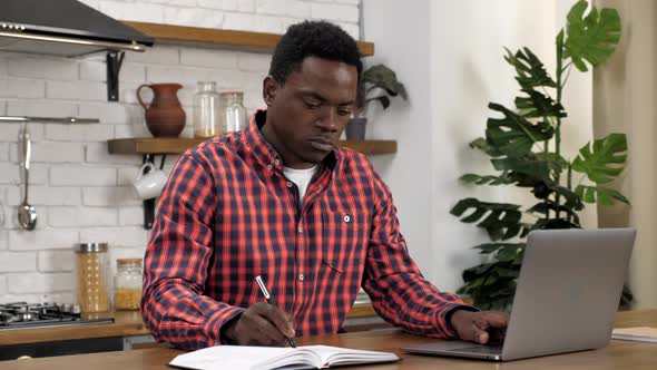 African American Man Freelancer Works for Laptop in Kitchen Writes in Notebook