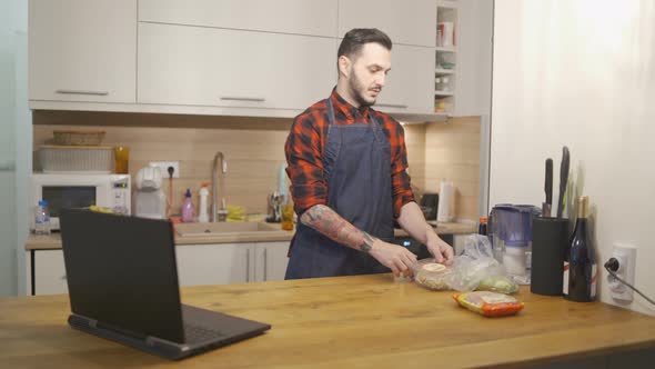 Young Man in Apron Moving Products From Kitchen Counter and Checking Laptop