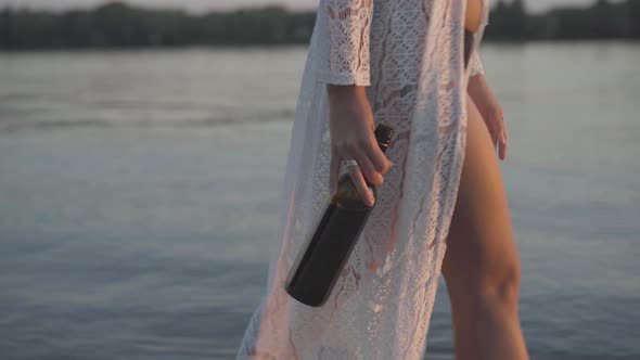 Unrecognizable Tanned Caucasian Woman in Swimsuit and Gown Walking with Wine Bottle Along River or