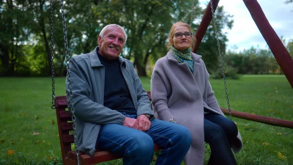 Happy Elderly Couple Talking And Relaxing On A Park Swing. Medium Shot