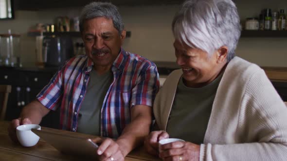 Happy senior mixed race couple having coffee laughing in kitchen