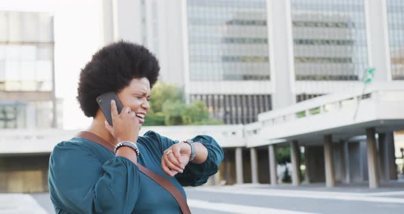 Happy plus size biracial woman talking on smartphone and checking smartwatch