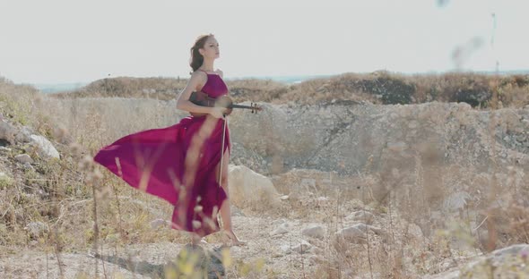 Woman in Windy Dress and Blowing Hair Stands with Violin and Bow on White Rock