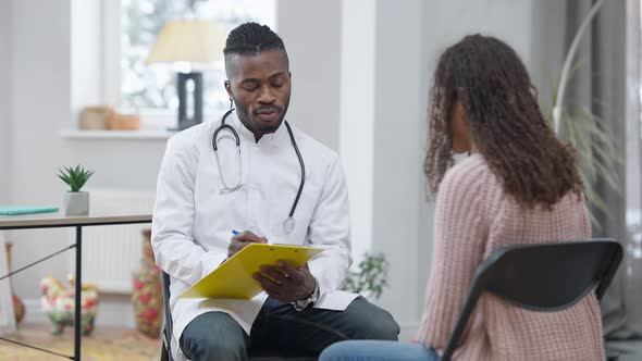 Portrait of Confident African American Doctor Writing Symptoms in Medical History Listening to Ill