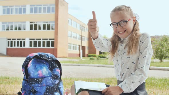 An Elevenyear Schoolgirl with a Book Sits on the Grass and Shows a Finger in Up