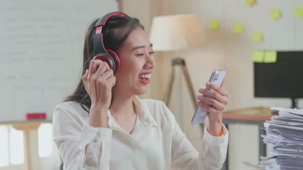 Close Up Of Asian Woman With Headphones Listening To Music On Smartphone After Working With Document