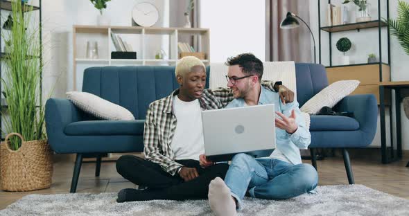 Mixed Race Gay Couple which Sitting on the Floor Near Sofa in Cozy Room and Use Laptop
