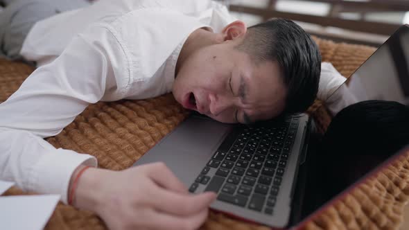 Tired Young Chinese Man Yawning Lying on Laptop Keyboard Napping in Home Office