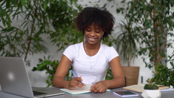 Cheerful African Female Student Wear Headphones Noting in Exercise Book Studying on Online Courses