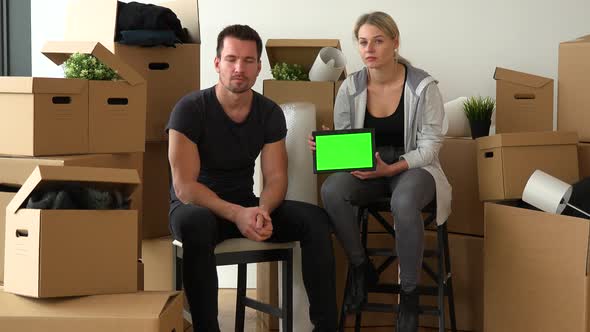 A Serious Moving Couple Sits on Chairs and Shows a Tablet with Green Screen To the Camera