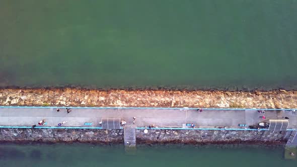 A dynamic top-down aerial shot of a breakwater jetty made out of stones with a paved pathway for civ