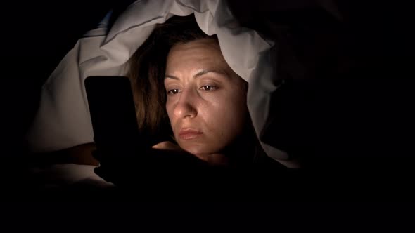 Young Woman in Bed with Smartphone. Woman Starring at Cellphone Device Before Going To Bed