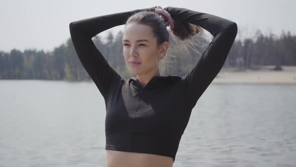 Attractive Slim Girl Straightens Her Horsetail, Standing with Arms Up in Front of the River