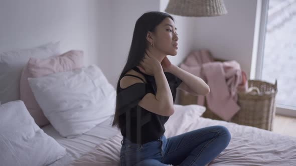 Tired Young Female Freelancer Stretching Neck Sitting on Bed Indoors