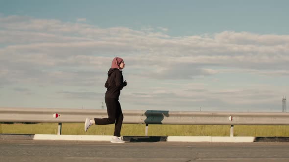 A Young Girl in a Black Tracksuit and Hijab, Running Along the Highway. Eastern Girl with a Scarf on