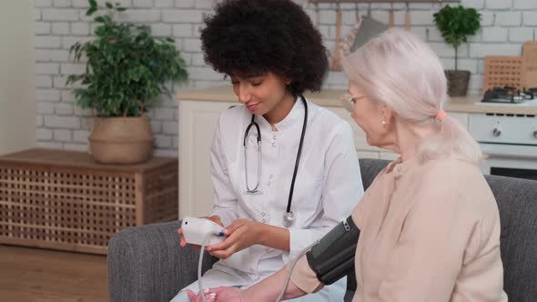 African American Woman Doctor Checking Blood Pressure of Senior Woman While Sitting on Couch at Home