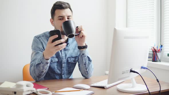 Young Office Worker Using His Phone at the Office Sitting at the Table with Computer Phone and