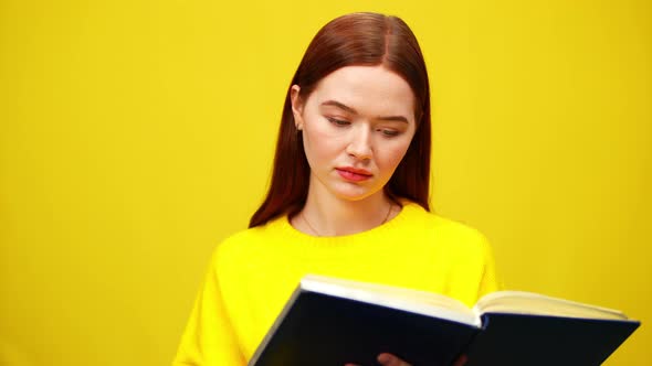 Concentrated Intelligent Millennial Woman Reading Book at Yellow Background and Smiling