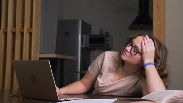 Woman with Glasses is Sitting at Desk and Working at Home She Bends Down and Holds Her Head She is