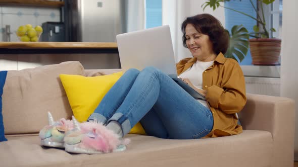 Happy Senior Woman in Unicorn Slippers Relaxing on Couch Using Laptop at Home