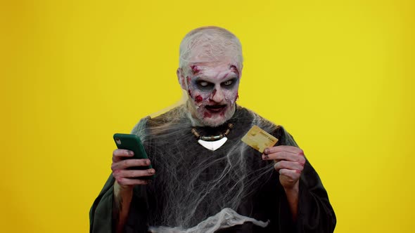 Sinister Man Halloween Zombie Using Credit Bank Card and Smartphone While Purchases Online Shopping