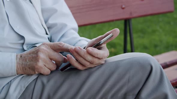 Close Old Senior's Hand Scrolling on Smartphone at Park's Bench