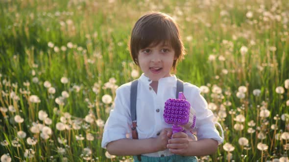 Portrait of a Five Year Old Boy in a Hat Stands on a Field of Dandelions and Shoots Soap Bubbles