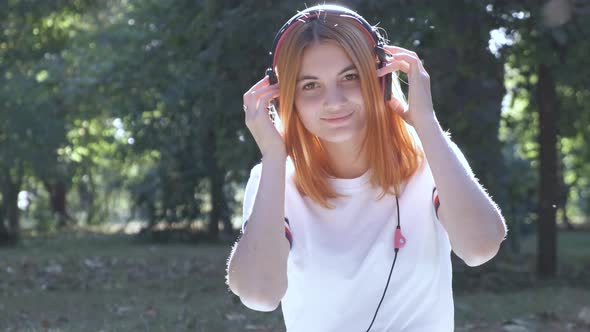 Pretty Teenage Girl with Red Hair Listening to Music in Earphones Outdoors in Summer Park