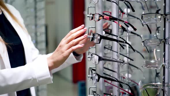 Customer choosing new glasses in the store of eyewear. Young woman turning stand with fashionable sp