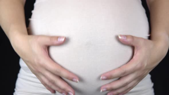 Pregnant Woman Stroking Her Stomach and Showing a Heart From Hands. Woman in a Light T-shirt