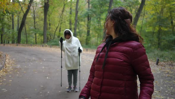 Smiling Mature Charming Woman Looking Back at Friend Endorsing Sportswoman and Strolling with Poles