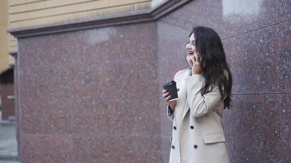 Beautiful Girl in Stylish Coat Talks Pleasantly on the Phone and Drinks Beverage