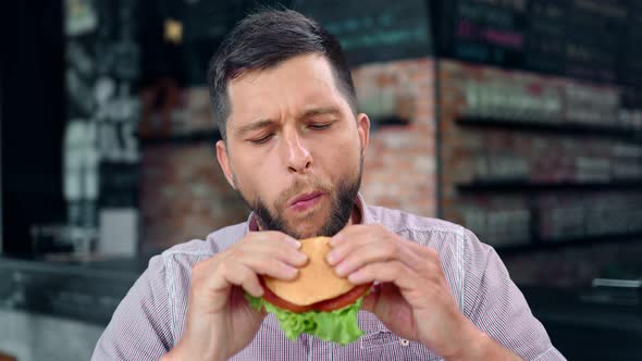 Hungry Bearded Young Slim Man Enjoying Biting Tasty Burger Feeling Pleasure Looking at Camera