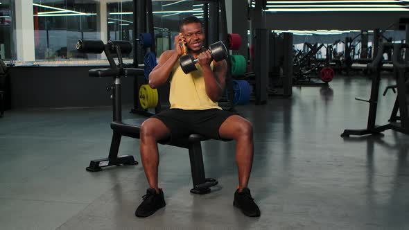 Strong Muscular African American Happily Talking on Mobile Phone in the Gym While Lifting Dumbbells