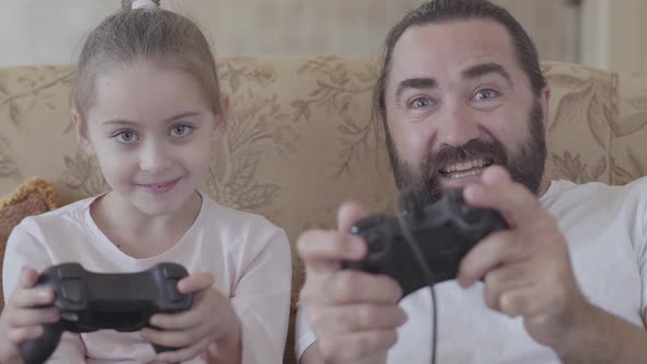 Bearded Man Sitting on the Sofa and Playing Video Game Holding Joystick with His Little Daughter