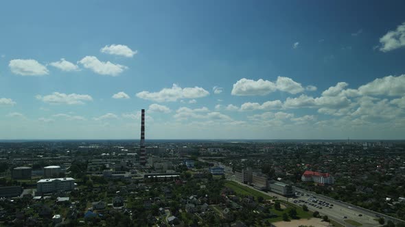 Flight Over The City. A Tall Factory Chimney Is Visible. In Backlit Sunlight. Aerial Photography.