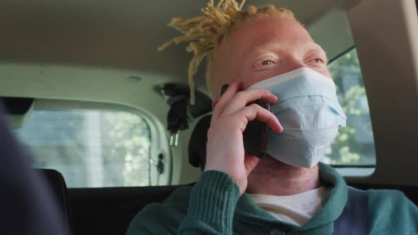 Happy albino african american man wearing face mask sitting in car talking on smartphone