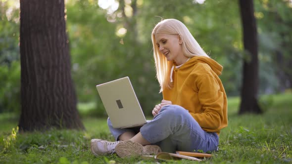 Young Blonde Woman Happily Talking By Video Call Using Laptop on Green Grass