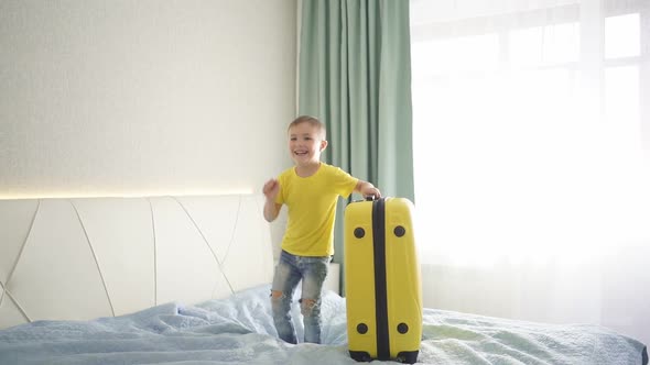 Happy Cute Boy Jumps on the Bed in Bedroom Near Packed Suitcase